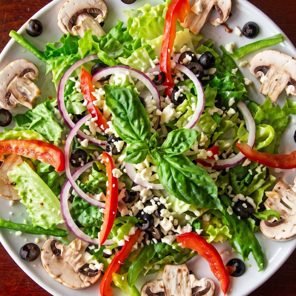 Overhead view of a colorful garden salad with lettuce, mushrooms, olives, peppers, red onion, feta, and basil on a white plate.